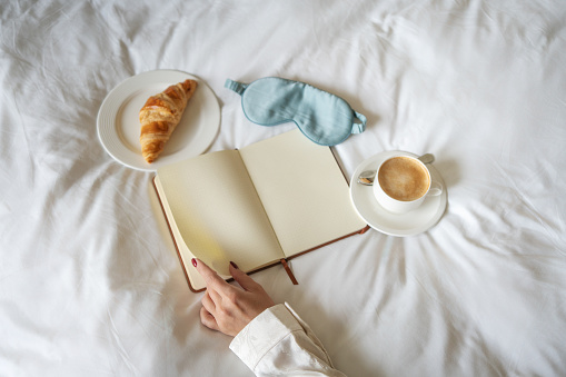 Woman's hand on an open journal beside a fresh croissant and cappuccino, with a sleep mask on white bed sheets. Morning journaling and diary concept.