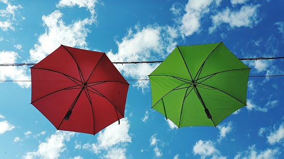 Two red and green umbrellas sway in the breeze, casting playful shadows on the sandy ground below. The clear blue sky and fluffy white clouds create a serene backdrop on a sunny day.
