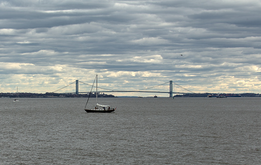 sailboat in new york harbor with verrazzano narrows bridge in the background (hudson river sunset with dramatic cloudy sky) suspension bridge small boat ship