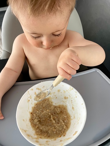A little boy, sitting on a high chair, eats porridge by himself with a spoon