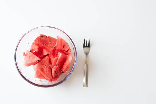 Sliced watermelon in a glass bowl with a fork, ready to eat sweet fruit. White background and copy space, flatlay.