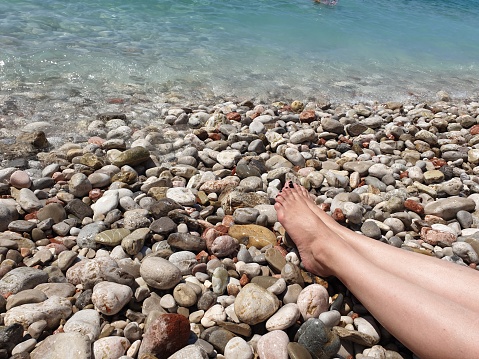 Woman resting legs and feet on a summer beach at the sea. Enjoying sun by sitting on a pebbles beach and watching blue sea water. Peaceful scene.