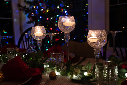 Christmas ornaments on a diner table with candles. Dark scene with a christmas tree blurred in the background.