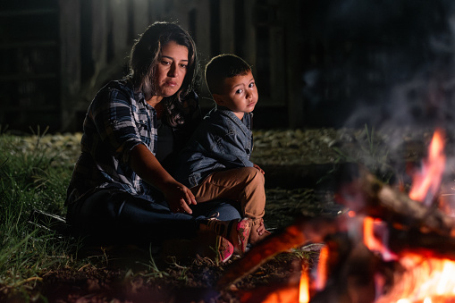 young latin woman carrying her son, sitting in front of a campfire at night, looking sad and bored.