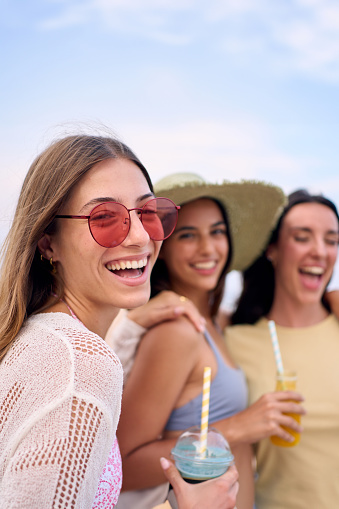 Vertical Portrait of cheerful three young Caucasian women posing smiling looking at camera on beach. Happy friends of generation z holding fruit smoothies enjoying summer holidays in Mediterranean sea