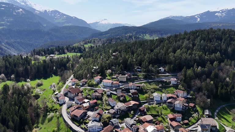 Idyllic small town on green mountain in Switzerland at sunlight. Snowy mountains of Alps in background. Aerial Forward flight.