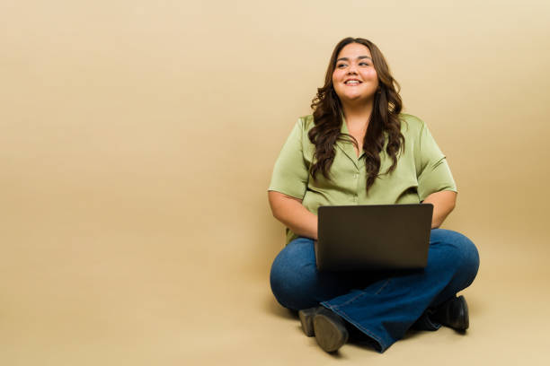istock Cute plus-size woman sits with a laptop on a beige studio backdrop 2149319197