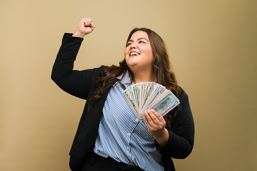 Triumphant plus-size woman cheerfully holds money in a studio, representing achievement