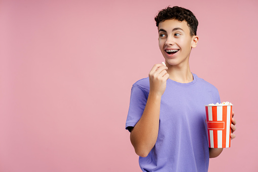 Smiling attractive teenager eating popcorn, holding bucket looking away at copy space standing isolated on pink background. Handsome boy with braces watching movie. Entertainment concept