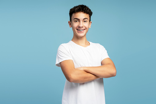 Handsome confident boy teenager with braces wearing casual white t shirt with crossed arms looking at camera standing isolated on blue background. Advertisement concept