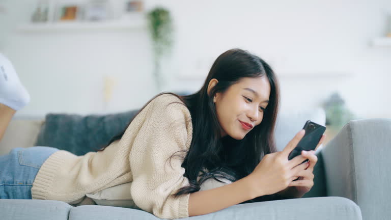 Happy young asian woman relax on comfortable couch at home texting messaging on smartphone, smiling girl use cellphone, browse wireless internet on gadget, shopping online from home