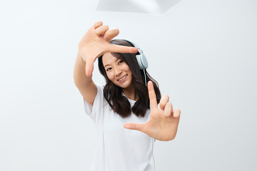 Happy smiling cute Asian young female in white basic t-shirt show frame shaped gesture at camera posing isolated on over white studio background. The best offer for ad. Favorite Music App Ad concept