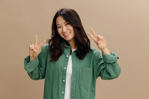 Lovely friendly Asian student young woman in khaki green shirt show v sign gesture smiling posing isolated on over beige pastel studio background. Cool fashion offer. Lifestyle and Emotions concept