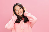 Smiling cutie cute Asian young lady in pink hoodie sweatshirt with cute headphones looks at you with love posing isolated on pink studio background. Good offer. Sound streaming platform ad concept
