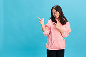 Cheerful Asian student young lady in pink hoodie sweatshirt wear round glasses point fingers aside posing isolated on over blue background. The best offer for ad. Eyewear for vision correction concept