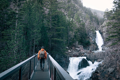 A man with an orange jacket and a backpack walks on an iron bridge. There is a waterfall near him. Concept of adventure and exploration. The man is hiking through nature.