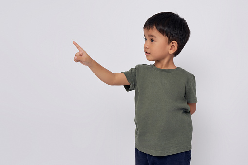 Smiling happy Asian boy 2-3 years old wearing a green t-shirt pointing finger at copy space isolated on white background