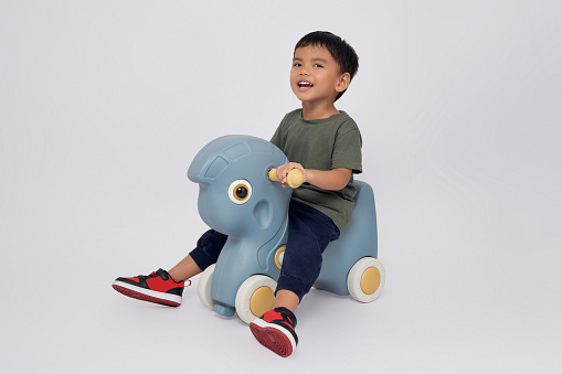 Full body little small smiling happy Asian boy 2-3 years old wearing green t-shirt  sitting on toy rocking horse, playing with a toy and looking at camera isolated on white background