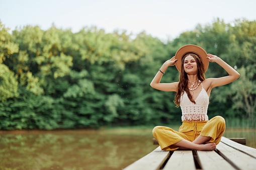 Hippie eco-activist woman traveler sits on a bridge by a lake with her arms outstretched with a hat and smiling sincerely. High quality photo