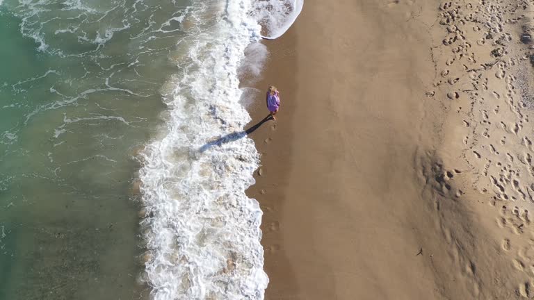 Aerial slow motion romantic young blonde woman in beautiful beach dress waving on wind walking by sandy beach, sea waves