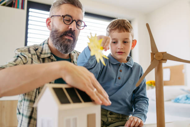 istock Father explaining renewable energy, teaching about sustainable lifestyle his young son. Playing with house with solar panels, wind turibine at home. 2149355665