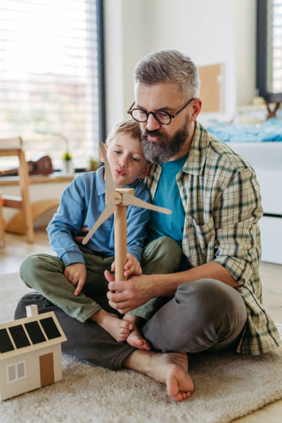 Father explaining renewable energy, teaching about sustainable lifestyle his young son. Playing with house with solar panels, wind turibine at home.
