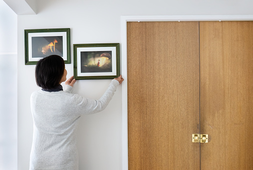 Woman hanging picture frames on a white wall at home. Fairytale glowing mushroom photos in the frames.