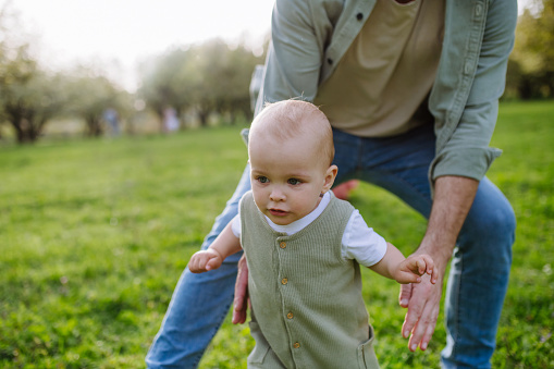 First steps for little toddler. A father supporting baby while walking in soft spring grass on meadow. Father's day concept.
