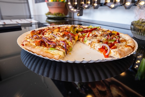 A Californiastyle pizza with pizza cheese and assorted ingredients sits on a paper plate on the table. One slice has been taken out of the fast food dish