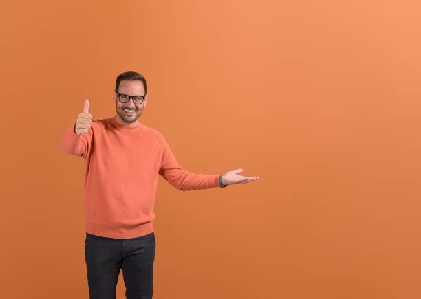 istock Happy businessman showing thumbs up sign and empty palm while sharing feedback on orange background 2149359184