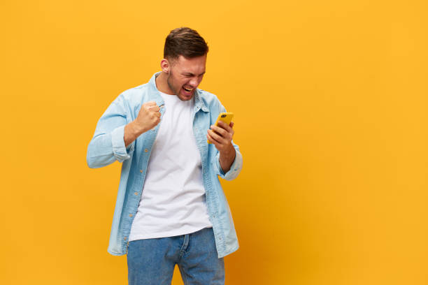istock Angry irritated tanned handsome man in blue basic t-shirt scream at smartphone posing isolated on orange yellow studio background. Copy space Banner Mockup. Online Social media concept. Bad news 2149361667
