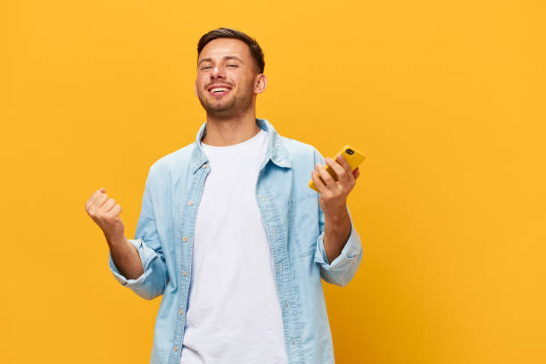 istock Smiling enjoyed tanned handsome man in blue basic t-shirt raise fist up using phone posing isolated on orange yellow studio background. Copy space Banner Mockup. Online Social media. Lucky win concept 2149361777