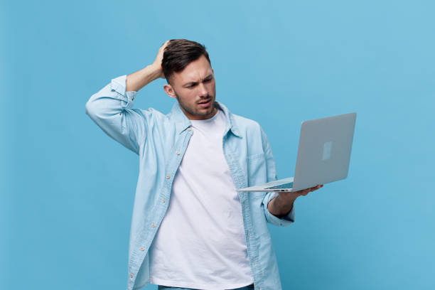 istock Confused tanned handsome man in casual basic t-shirt touch head have problem with broken laptop posing isolated on blue studio background. Copy space Banner Mockup. Electronics repair IT concept 2149362182