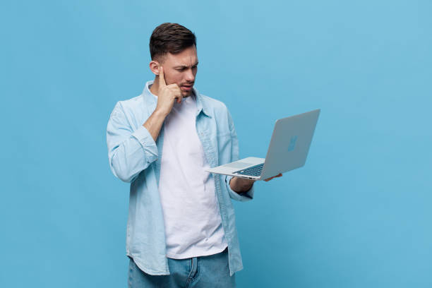 istock Thoughtful tanned handsome man in casual basic t-shirt concerned make difficult decision with laptop posing isolated on blue studio background. Copy space Banner Mockup. Electronics repair IT concept 2149362349