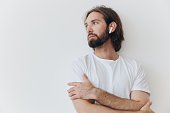 A man with a beard and long hair in a white T-shirt and blue jeans stands against a white wall, leaning against it and listening to music with wireless white headphones, staring thoughtfully