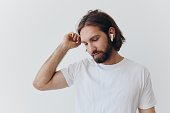 A man with a beard and long hair in a white T-shirt and blue jeans stands against a white wall, leaning against it and listening to music with wireless white headphones, staring thoughtfully