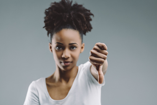 Afro-haired woman gives thumbs-down gesture, with a discerning look, set against a plain background