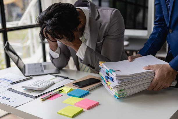 Male accountant stressed with too much paperwork and exhausted from work, covering his face with friend holding a lot of documents.