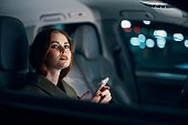 a close horizontal portrait of a stylish, luxurious woman in a leather coat sitting in a black car at night on the passenger seat, thoughtfully looking out the open window of the car
