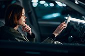 horizontal portrait of a stylish, luxurious woman in a green leather coat, sitting in a black car at night on the passenger seat, thoughtfully looking at the road stretching her hand forward