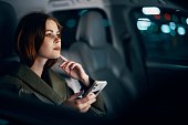 stylish, luxurious woman in a leather coat sitting in a black car at night on the passenger seat, looking thoughtfully to the side, holding her hand near her face, and the other holding a smartphone