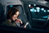 a close horizontal portrait of a stylish, luxurious woman in a leather coat sitting in a black car at night in the passenger seat, thoughtfully looking at her smartphone with her hand near her face