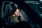 horizontal portrait of a stylish, luxurious woman in a green leather coat sitting in a black car at night on the passenger seat, thoughtfully looking at the road holding her hand near her face