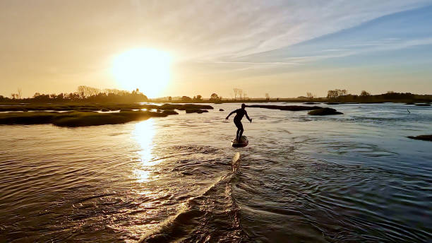 Mature surfer in wetsuit with surfboard on the beach, prepares to surf at night illuminated by his electric car headlights. Health, wellbeing & adventure sport.
