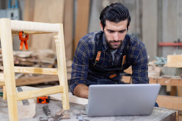 istock young carpenter caucasian man with laptop make drawing on wood to design at workspace. craftsman profession in wood factory 2149374978