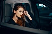 horizontal photo from the side, at night, of a woman sitting in a black car and thoughtfully looking forward holding her hand near her face