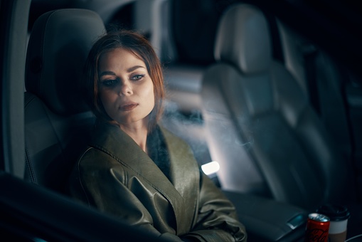 a close horizontal portrait of a stylish, luxurious woman in a leather coat sitting in a black car at night in the passenger seat, looking to the side through the fogged glass. High quality photo