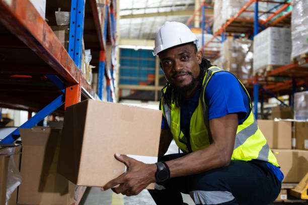 istock Man american african professional worker wearing safety uniform and hard hat holding box product on shelves in warehouse. Man worker check stock inspecting in storage logistic factory. 2149376780