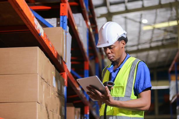 istock Man american african professional worker wearing safety uniform and hard hat using digital tablet inspect product on shelves in warehouse. Man worker check stock inspecting in storage logistic factory. 2149376983