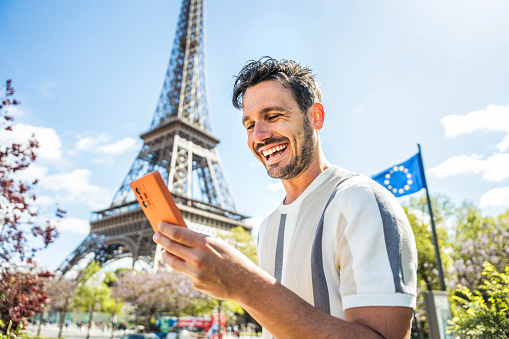 Happy tourist booking hotel with smart mobile phone device in Paris, France - Handsome young man using date app with smartphone - Travel, technology and youth concept